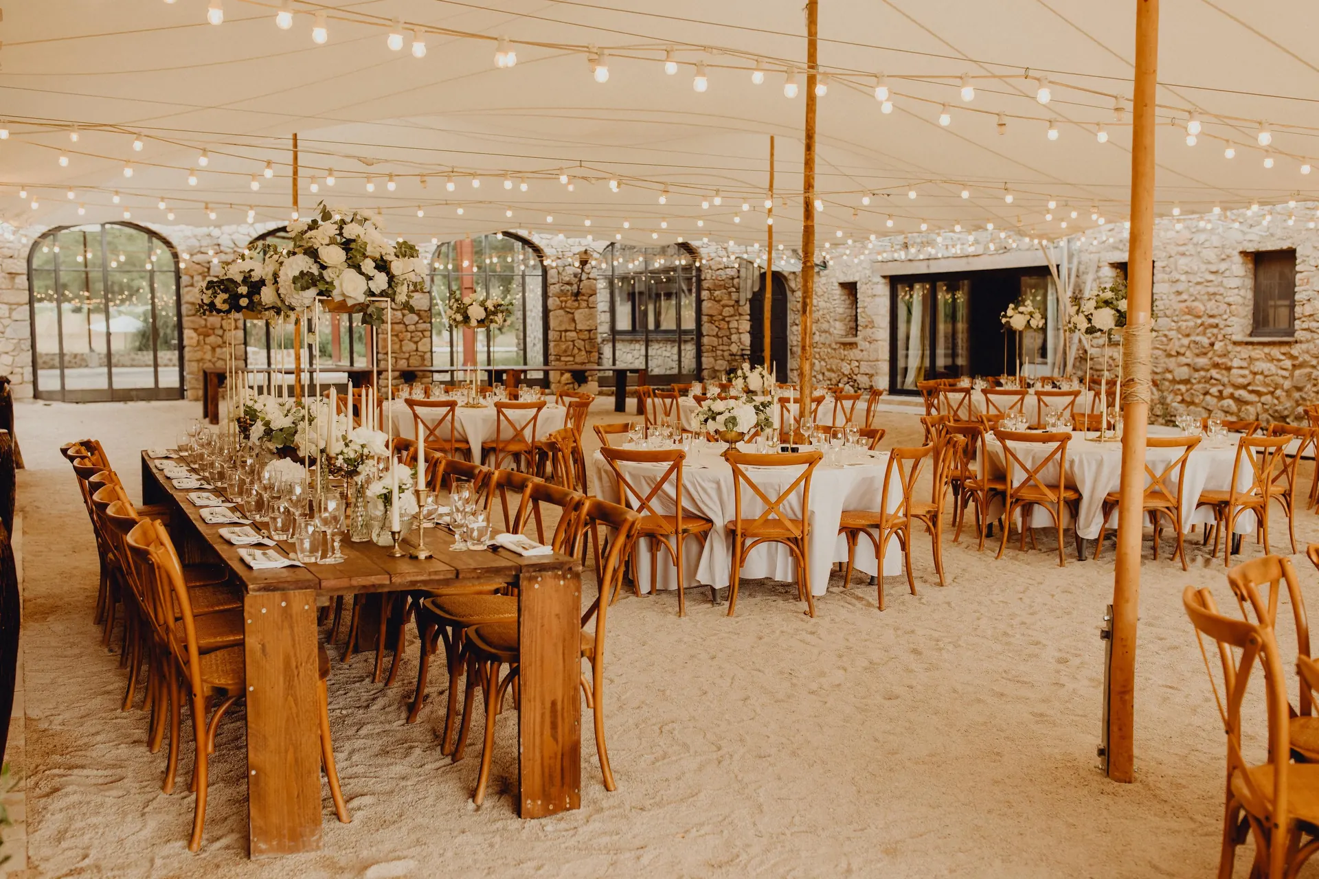 Réception dressée sous une tente illuminée de guirlandes : tables en bois, chaises croisées et compositions florales